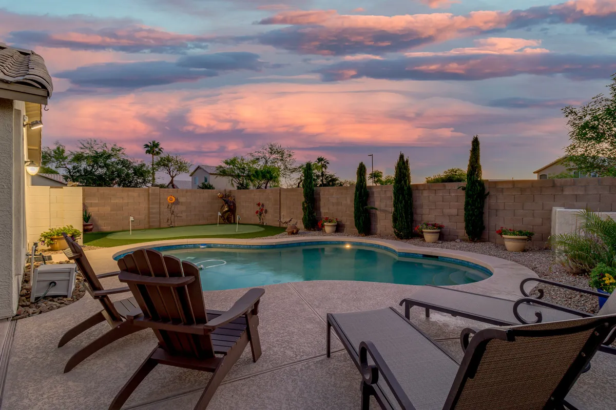 A serene backyard scene at sunset, featuring a sparkling swimming pool, plush lounge chairs, and a well-manicured green grass area. The sky glows with warm evening colors, inviting relaxation and gatherings by the water's edge.