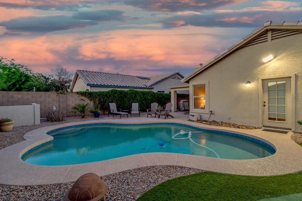 A peaceful backyard oasis with a sparkling swimming pool, comfortable lounge chairs, vibrant greenery, and a dramatic sunset sky. The setting is ideal for swimming, entertaining, or enjoying tranquil evenings outdoors.