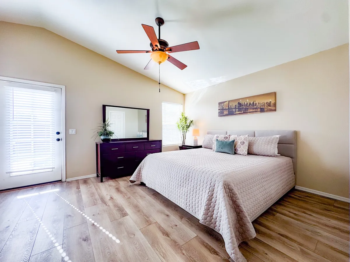 A tranquil bedroom featuring a plush bed adorned with textured bedding and decorative pillows. A dresser with a mirror, a window with blinds softly diffusing natural light, and a potted plant add to the peaceful ambiance. The overall scene exudes comfort and relaxation, perfect for restful nights and serene mornings.