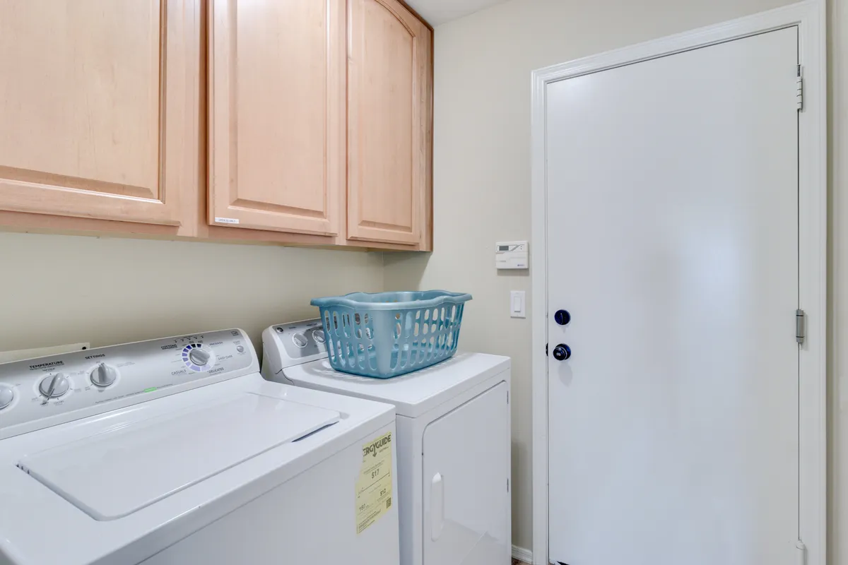 A tidy laundry space featuring a washing machine and dryer, overhead cabinets for storage, and a laundry basket. The clean, organized design highlights convenience for daily living and longer stays.