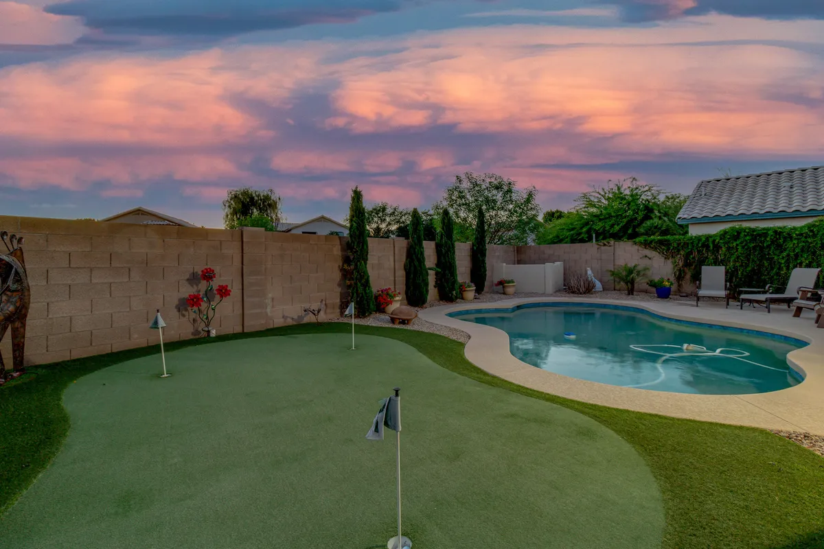 A vibrant backyard scene showcasing the putting green, swimming pool, flowering planters, and tall cypress trees. Outdoor seating is visible, and the lush landscaping creates a sense of privacy and abundance. The setting sun casts a golden glow, enhancing the tranquil vibe.