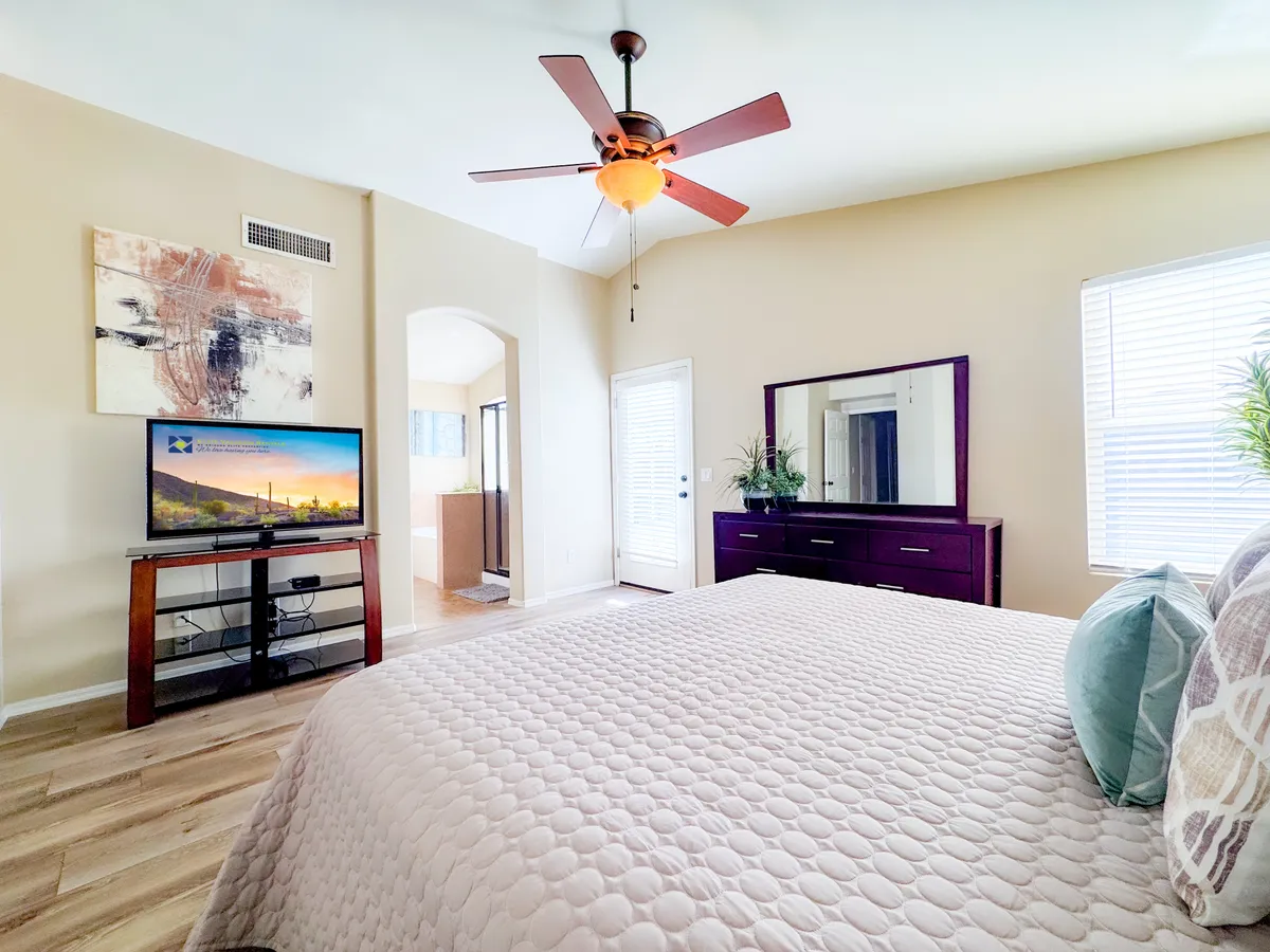 A restful bedroom with a cozy bed featuring a textured quilt, ceiling fan, wall-mounted television, and artistic wall decor. A dressing table with a mirror sits nearby, and natural light streams in from the window, creating a peaceful and home-like atmosphere.