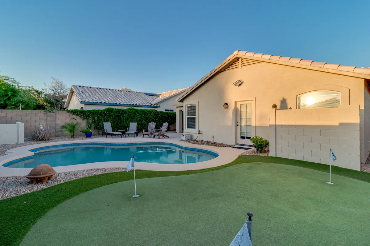 A vibrant, wide-angle view of the backyard featuring a shimmering pool, a golf putting green, lounge chairs, and the house facade. The scene is bathed in natural light, suggesting the enjoyment of both daytime and evening activities in a serene environment.
