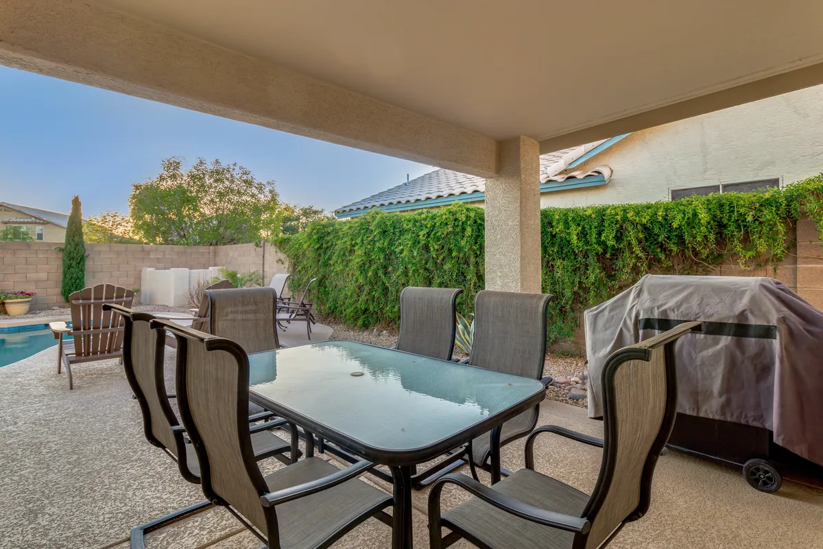 A cozy, well-furnished outdoor dining area set beside the pool, with a grill visible and landscaped garden creating a lush backdrop. The scene is staged for alfresco dining and entertaining, with evening light adding warmth to the atmosphere.