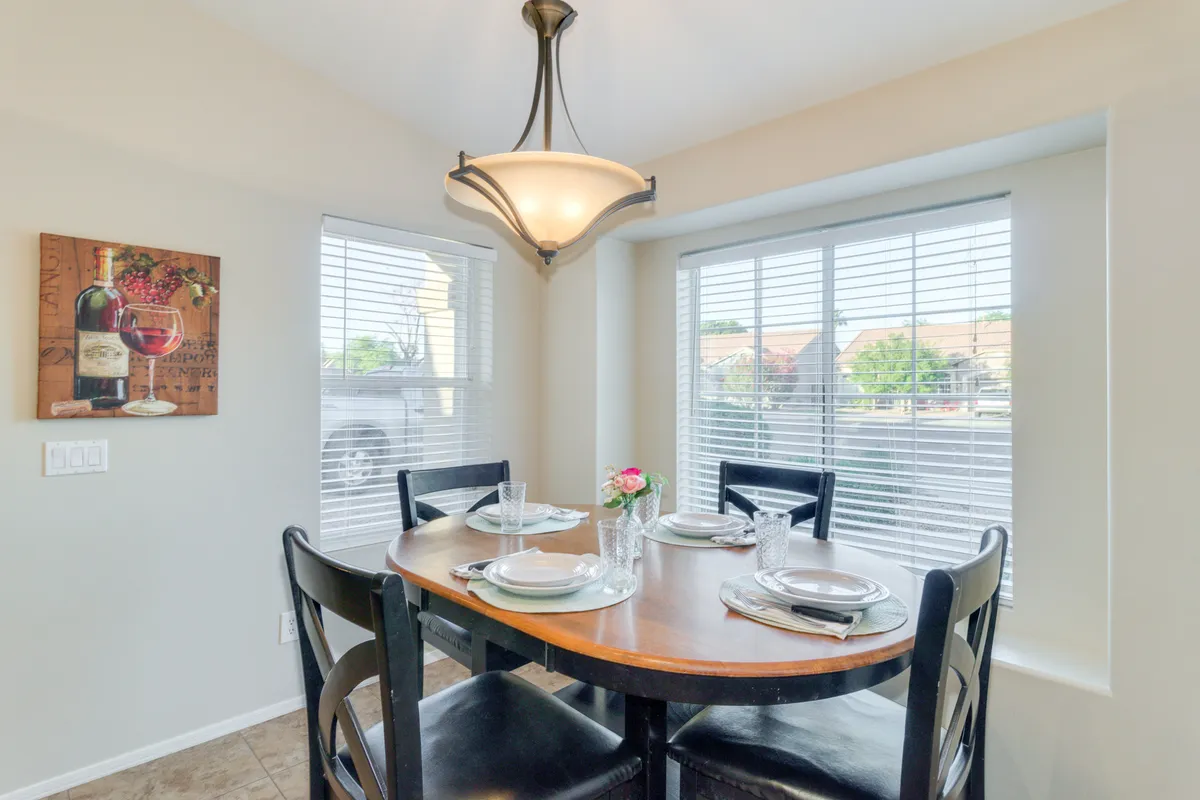 A cozy dining nook with a round wooden table, decorative wall art featuring wine, a floral centerpiece, and ample natural light streaming in through the windows. The setting is perfect for intimate family breakfasts or memorable gatherings.
