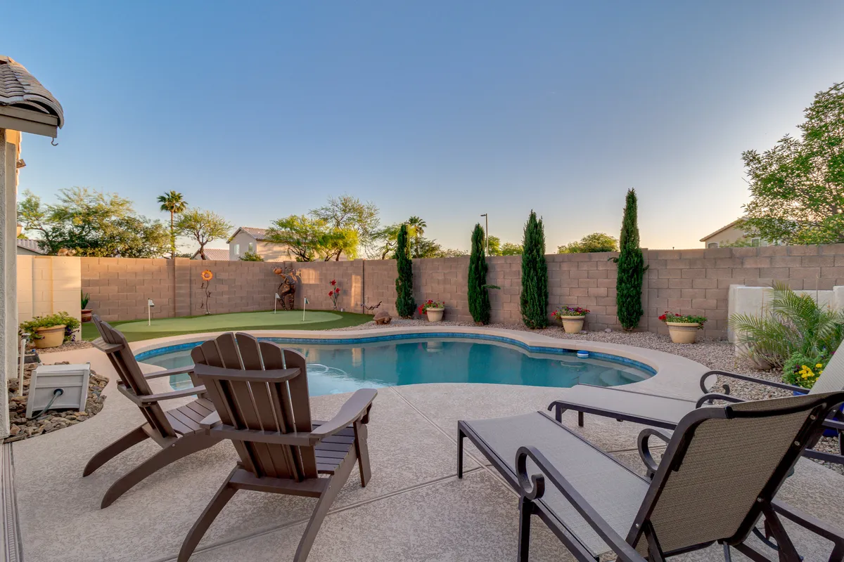 A tranquil backyard view featuring a swimming pool, adirondack chairs for lounging, lush landscaping, and tall cypress trees. The image captures a peaceful yet inviting atmosphere, ideal for family gatherings, sunbathing, or relaxing with a book.
