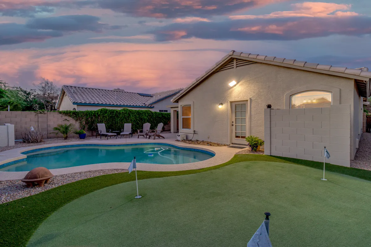 A wide-angle exterior shot of the backyard featuring a sparkling swimming pool in the foreground, a well-maintained putting green to one side, and a spacious patio area with seating. Lush landscaping surrounds the scene, creating a private and tranquil atmosphere ideal for relaxation and entertaining guests.